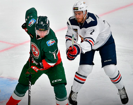 На изображении запечатлен момент хоккейного матча. Игрок в зеленой форме с шайбой противостоит сопернику в бело-синей форме. Оба игрока борются, сосредоточены на игре. Виден фрагмент ледовой арены