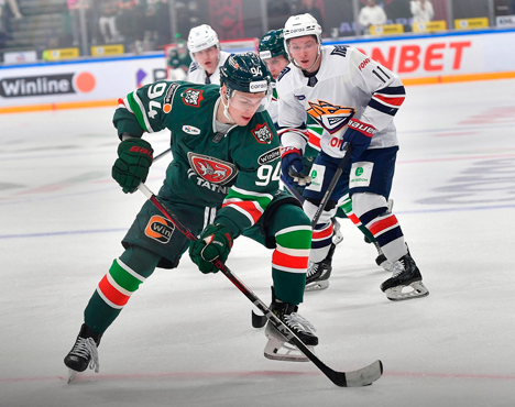 Хоккеисты в форме двух команд соревнуются на ледовой арене. Игрок в зелёной форме с клюшкой контролирует шайбу, игроки в бело-синей форме пытаются отобрать её. На заднем плане видны рекламные баннеры и зрители